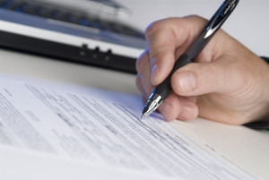 man's hand holding a pen writing a signature, computer in background